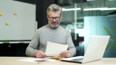 Smiling mature gray haired bearded man in glasses reading letter with good news while sitting at desk at workplace in modern office. A happy middle aged worker is contentedly reviewing a document