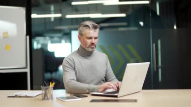 Serious mature gray haired bearded businessman is typing on a laptop while sitting at a desk at a workplace in a modern office Confident concentrated middle aged entrepreneur works online Writes email