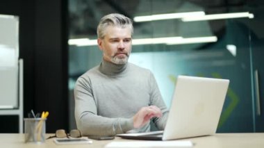 Happy mature gray-haired bearded businessman typing on laptop while sitting at desk at workplace in modern office. Confident friendly middle aged entrepreneur smiling and posing looking at the camera