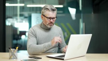 Overworked mature gray haired bearded businessman in glasses tired working on laptop at desk at workplace in modern office. The entrepreneur takes off his glasses and massages the bridge of his nose