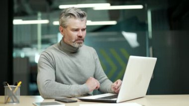 Confused mature gray haired bearded businessman is solving a complex problem while working on a laptop while sitting at a desk in modern office. Disappointed entrepreneur can't find the right solution