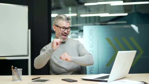 快乐而成熟的白发商人戴着眼镜 高兴地在办公桌前跳舞 看着办公室里的笔记本电脑屏幕 笑着兴奋的企业家庆祝商业成功 — 图库视频影像