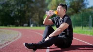 Happy young adult asian sports man drinking water from a bottle while sitting on a treadmill in a city city stadium. A toned handsome man in a sports suit is smiling and relaxing after training