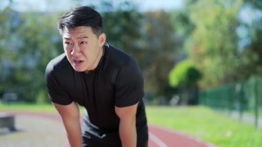An adult asian runner is short of breath after a fast sprint run on a treadmill in an urban city stadium. An athlete in a sports suit leans on his knees and breathes deeply. Sport and training concept