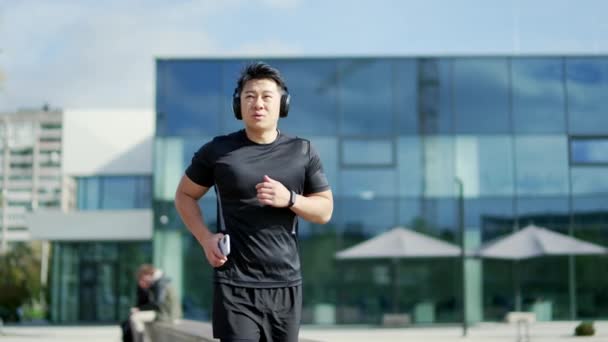 現代的な都市の建物を背景に 路上でヘッドフォンやスマートフォンのジョギングをしている若い大人のアジアのランナー トラックスーツの男は音楽を聴きながら外でジョギングを楽しむ — ストック動画