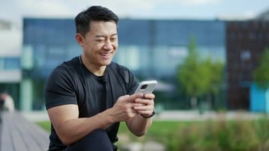 Smiling adult asian man using a smartphone while sitting on a bench against the background of a modern city building. A handsome male is reading a pleasant message, texting with a friend or girlfriend