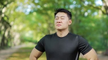 Close up of a young adult asian sports man relaxing with his eyes closed after a morning run while standing in an urban city park. A handsome male breathes calmly and enjoys a beautiful sunny day