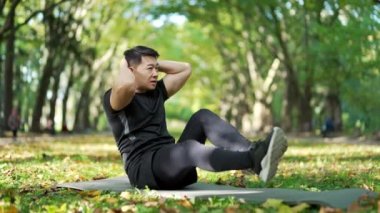 Asian man exercises abdominal muscles while sitting on a mat in an urban city park. Handsome male in a sports suit is engaged in gymnastics. Healthy and young athlete busy with training and exercise