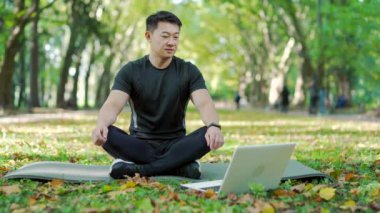 Asian man in tracksuit doing yoga on mat in urban city park watching online class on laptop. Male meditating outdoors in lotus position with eyes closed and taking slow deep breaths with calmness