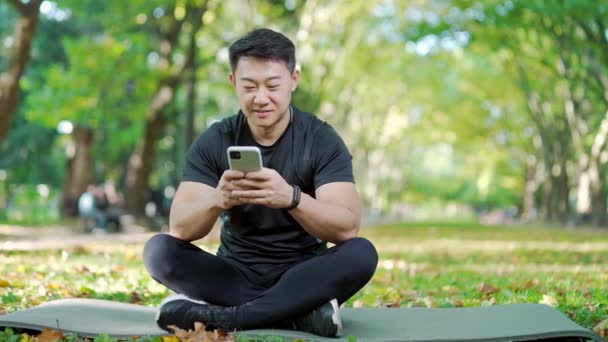 Feliz Hombre Asiático Chándal Sentado Estera Teléfono Inteligente Navegación Parque — Vídeo de stock