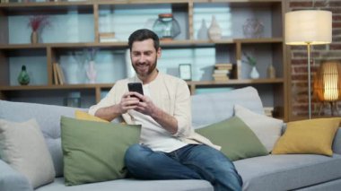 Gülümseyen sakallı, olgun bir adam akıllı telefonu tutuyor ve evde yalnız başına sosyal medya izliyor. Yakışıklı bir erkek, oturma odasında kanepede oturmuş, internette internet ürünlerini karıştırıyor. 