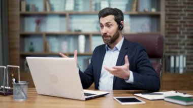 Smiling mature business man investor with wireless headset looking at the camera and talking by online meeting conference video call at modern office workplace indoors Working conversation concept