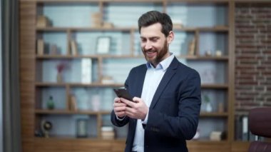Gülümseyen iş adamı yatırımcı akıllı telefonu tutar ve modern ofis işyerinde sosyal medyayı izler Mutlu sakallı girişimci işvereni telefon uygulaması kullanarak evde tek başına dolaşır. 