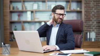 Overworked tired mature business man investor with glasses suffers with severe pain in muscles of the neck shoulder from hard work on computer at desk in the modern workplace Osteochondrosis concept