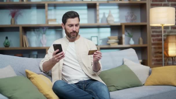 英俊而成熟的男人坐在客厅沙发上 一边在智能手机上输入信用卡号码 一边在网上购物 一边在家里收钱 快乐的顾客 — 图库视频影像