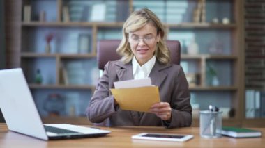 Happy mature businesswoman reading letter with good news while sitting at desk at workplace in office. A smiling woman in a suit and glasses is satisfied with the contents of the received document