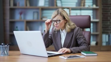 Shocked mature businesswoman looking surprised at laptop screen while sitting at desk at workplace in office. Sad woman holds her head with her hands because she saw unpleasant content, read bad news