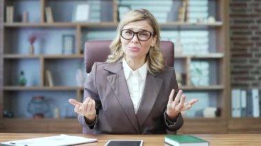 Webcam view. Angry mature businesswoman arguing with subordinate looking at camera while sitting at desk in office. Annoyed woman in a suit and glasses talks about problems gesturing with her hands