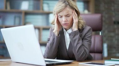 Tired mature businesswoman feeling headache while working on laptop at workplace at desk in office. A sad and overworked woman in a suit massages her temples and rubs a sore spot with her hands