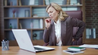 Tired mature businesswoman due to sedentary lifestyle feels back pain while working on laptop at workplace at desk in office. Sad woman in a suit is massaging and rubbing her lower back with her hands