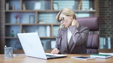 Tired mature businesswoman feeling neck pain while working on laptop at workplace at desk in office. A sad and overworked woman in a suit massages her muscles and rubs a sore spot with her hands