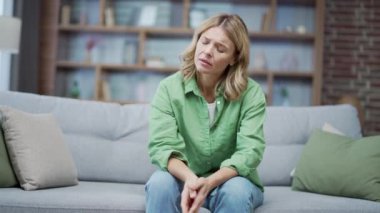 Mature blonde woman has a bad mood sitting on the sofa in the living room at home. Upset woman in casual clothes thinking about her problems indoors. She is tired, unhappy, disappointed and depressed