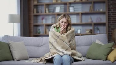 Blonde mature woman is frozen sitting in cold room on sofa at home. A disgruntled female in casual clothes is wrapped in a blanket and warms her hands by rubbing them. Heating problem concept