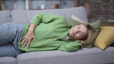 Sick mature blonde woman feeling stomach ache while lying on sofa in living room at home. Tired female on the couch has an upset stomach, acute gastritis or menstruation, feeling of spasms, heartburn