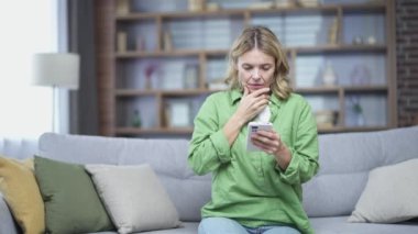 Sad blonde mature woman read bad news on smartphone while sitting on sofa in living room at home. Emotional female holding hand on head, worried, thinking about tragic event, excited about problem