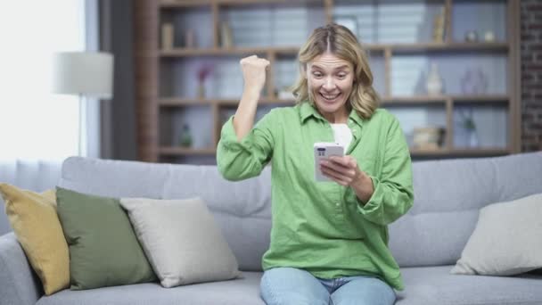 Happy Mature Blonde Woman Joyfully Browsing Smartphone While Sitting Sofa — Stockvideo
