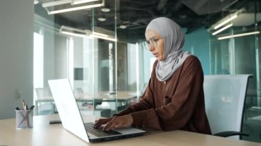 Soğuk ve genç Müslüman iş kadını gözlüklü tesettüre giriyor ve titriyor. Elleri titriyor. Modern ofisteki masadaki dizüstü bilgisayarda çalışmaya çalışıyor.