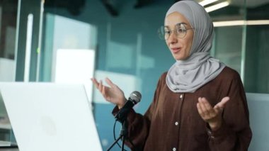Muslim businesswoman in hijab doing video meeting consultation conference podcast at modern office Lecturer teacher talking into microphone while recording content for online education course indoors