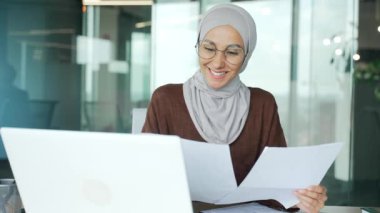 Smiling young muslim businesswoman in hijab looking on business documents at desk in modern office Happy entrepreneur with glasses have paper work and typing browsing on laptop computer at workplace 