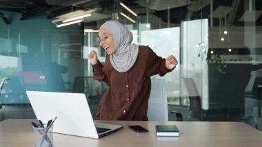 Tesettürlü genç Müslüman iş kadını dizüstü bilgisayarından memnun. İyi iş çıkarmışsın ve ofiste dans ediyorsun. İyi çalışmalar, iyi çalışmalar, iş yerindeki bilgisayar projelerini bitirdikten sonra rahatla.