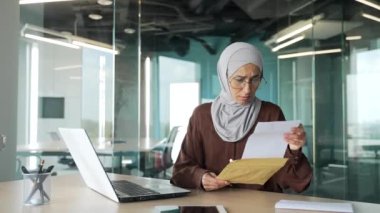 Sinirli, üzgün, gözlüklü, tesettürlü genç Müslüman iş kadını, modern işyerinde maaş indirimi veya işten çıkarma kararında kötü haber içeren açık bir mektup aldı.