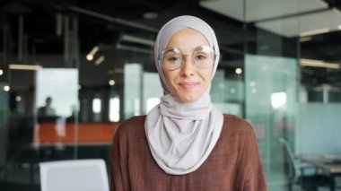 Close up portrait of smiling young muslim businesswoman employee in hijab looking at the camera in glass modern workplace Happy entrepreneur with glasses enjoying great workday in the office indoors