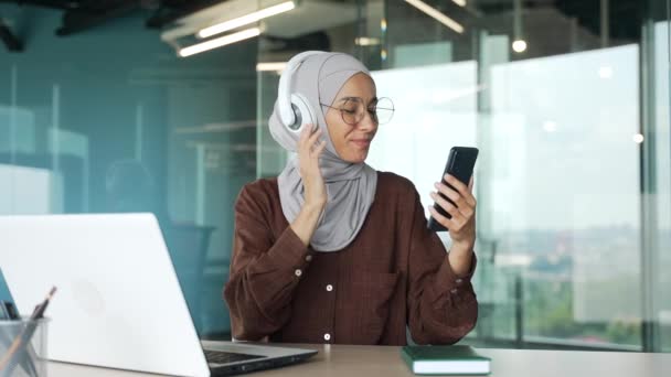 ヘッドフォン付きヒジャーブ州の陽気なイスラム教徒の若い実業家は スマートフォンを保持し ガラスのオフィスで音楽ダンスを聴く現代の職場でリラックスして楽しんで眼鏡をかけた起業家を笑顔 — ストック動画