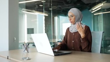 Muslim young businesswoman entrepreneur in hijab feels hot during computer work and turn on remote control air conditioner cooling system or humidifier changing temperature at modern office workplace