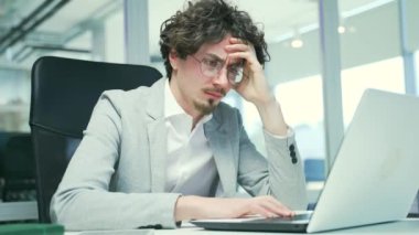 Thoughtful busy young business man with glasses typing on laptop computer working online at desktop in modern office Concentrated bearded entrepreneur takes responsible approach to the project indoors