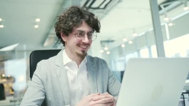 Friendly young bearded business man employee with glasses looking at the camera and talking by online meeting conference video call on laptop at modern workplace indoors Working conversation concept