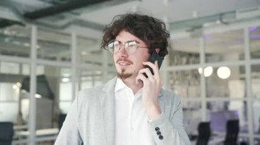 Smiling curly haired business man entrepreneur talking on mobile phone at modern office indoors Successful busy bearded office worker having professional conversation chat on smartphone at workplace