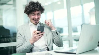 Overjoyed business man freelancer extremely happy after read message on smartphone at modern workplace Excited entrepreneur look at phone screen and pleased rejoices received great news at office desk