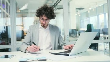Focused busy young business man with glasses typing on laptop computer working online at desktop in modern office Concentrated bearded freelancer make notes in working notebook to the project indoors