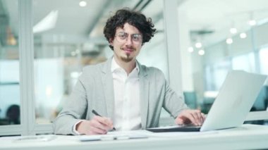 Portrait of smiling bearded businessman with glasses looking at the camera at modern glass office workplace Successful curly entrepreneur creative freelancer in formal suit sitting at desktop indoors