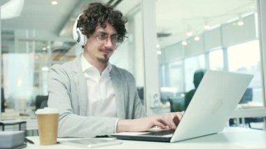 Gülümseyen genç iş adamı, kulaklıklarıyla bilgisayarda yazı yazıyor, bilgisayar başında müzik dinliyor, modern ofis, sakallı, mutlu girişimci, kapalı alanda iyi iş gününün keyfini çıkarıyor.