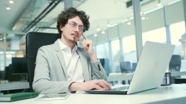 Thoughtful young curly haired business man freelancer with glasses reflects on the current project considers options typing on laptop computer working online at desktop in glass modern office indoors