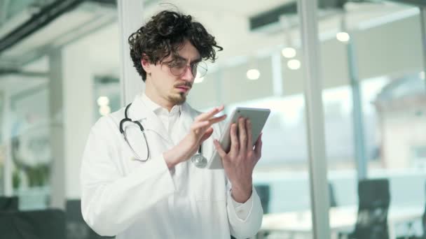 带听诊器身穿白色长袍的卷曲年轻医生在医院诊所拿着数码平板电脑成功的医生戴着眼镜在室内用小玩意发短信 — 图库视频影像
