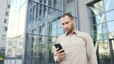 Yoğunlaşmış genç işadamı, bir ofis binasının yanında caddede dururken akıllı telefon kullanıyor. Ciddi bir erkek elinde telefon tutuyor, e-postaları kontrol ediyor, bir müşteriye mesaj yazıyor.