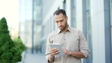 Ofis binasının önünde dikilirken tablet kullanan, gömlekli, genç ve ciddi bir iş adamı. Yakışıklı, melez bir erkek, iş günü boyunca internette internetten bilgi topluyor.