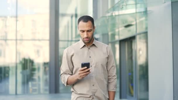 一位年轻人在一幢办公楼前散步时 一边看手机一边用手机发短信一位男性在用智能手机发短信 — 图库视频影像
